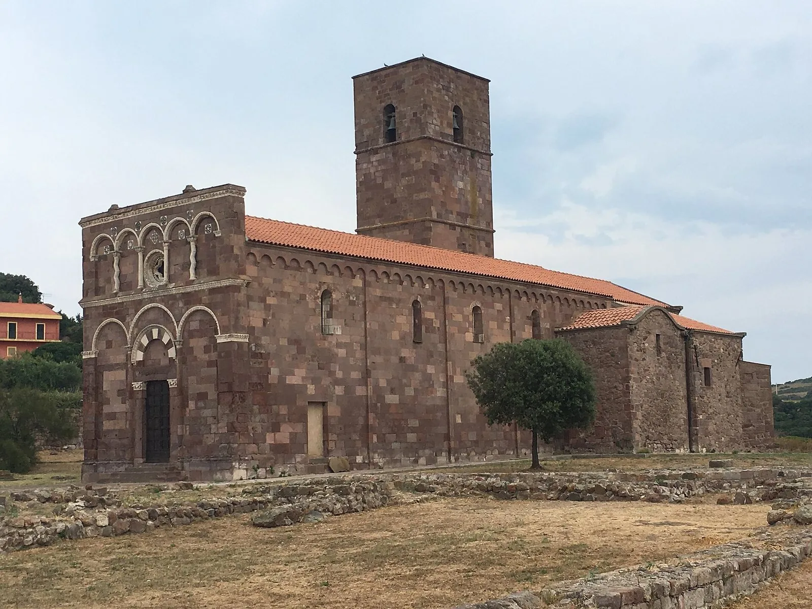 La chiesa romanica di Nostra Signora di Tergu, in pietra rossa con facciata ad archetti e campanile quadrato, vista di tre quarti tra i resti del monastero