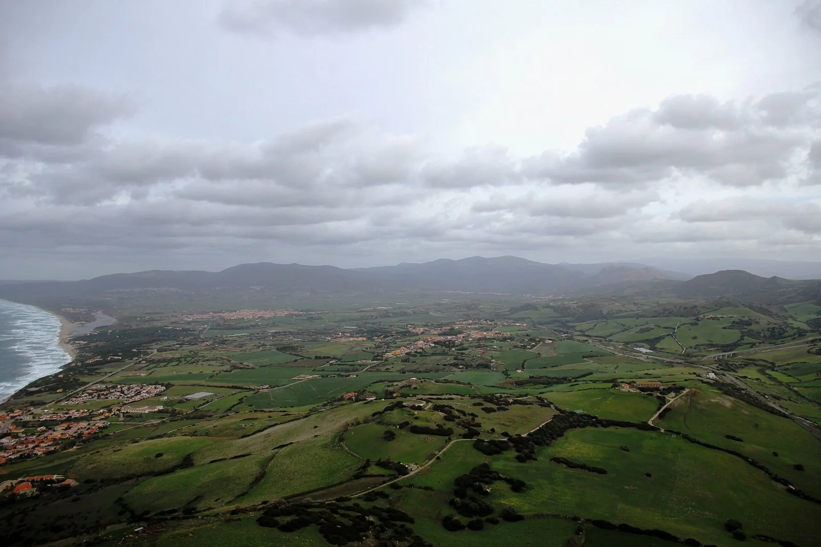 Panorama dell'Anglona dall'alto: campi verdi, paesi sparsi e strade di campagna, con la costa e una lunga spiaggia sulla sinistra sotto un cielo nuvoloso