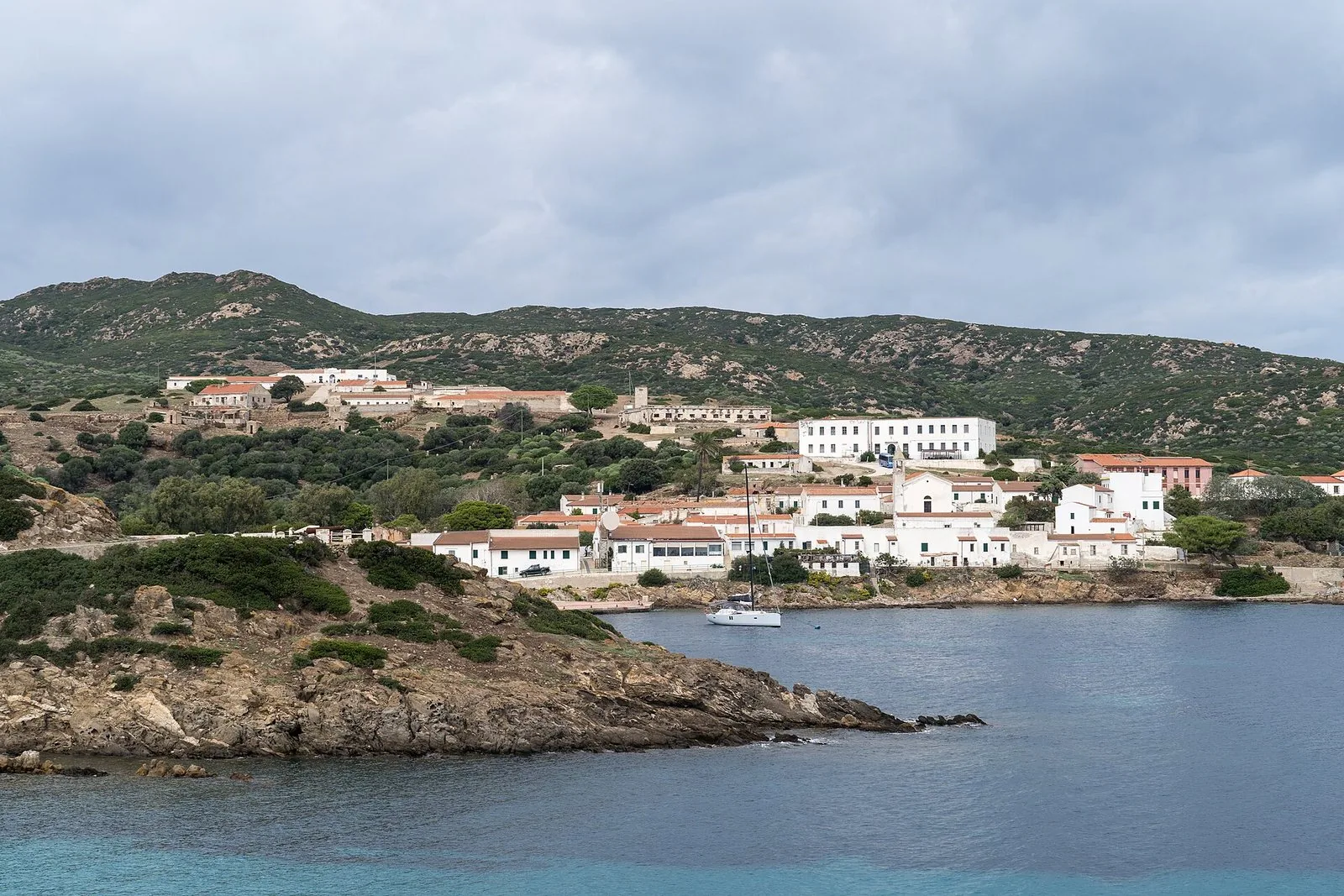 Il borgo di Cala d'Oliva all'Asinara: case basse bianche con tetti rossi sulla collina di macchia mediterranea, affacciate su una cala con una barca a vela